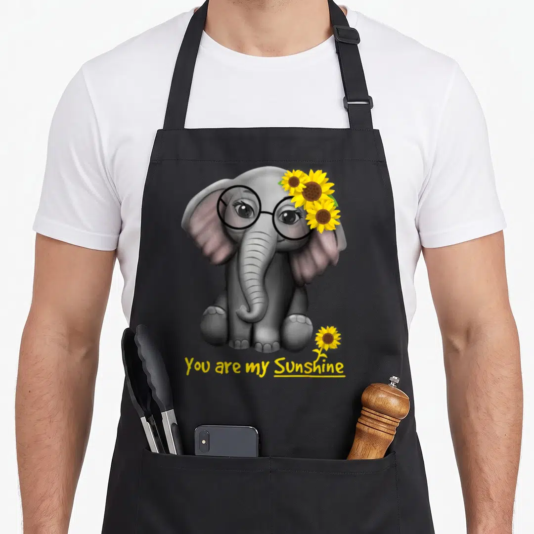 Clothe sunflower apron - Grey elephant wearing glasses with yellow sunflowers and You are my Sunshine text — shown on a black cotton canvas apron against a white background