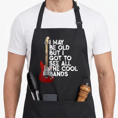 See but cooler apron - Red electric guitar with tan neck beside seven lines of distressed white text reading I MAY BE — shown on a black cotton canvas apron against a white background