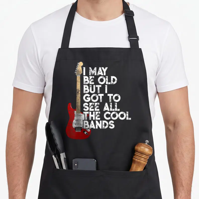 See but cooler apron - Red electric guitar with tan neck beside seven lines of distressed white text reading I MAY BE — shown on a black cotton canvas apron against a white background