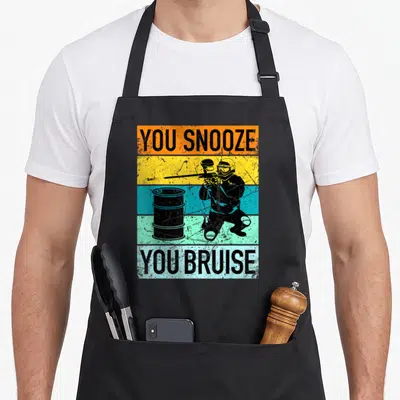 Funny gear paintball apron - You Snooze You Bruise text flanking a crouching paintball player behind a barrel on stripes — shown on a black cotton canvas apron against a white background