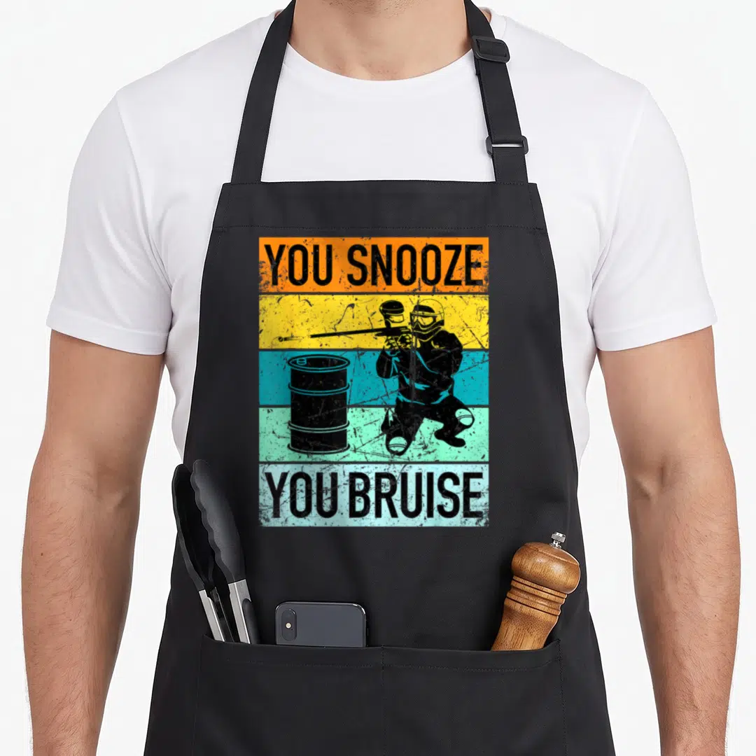 Funny gear paintball apron - You Snooze You Bruise text flanking a crouching paintball player behind a barrel on stripes — shown on a black cotton canvas apron against a white background
