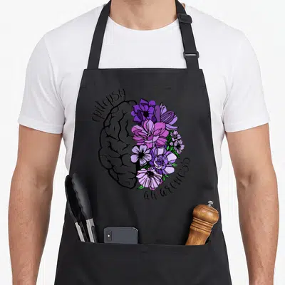 Flower epilepsy awareness apron - Split brain illustration with grey line art left side and purple pink flowers right — shown on a black cotton canvas apron against a white background