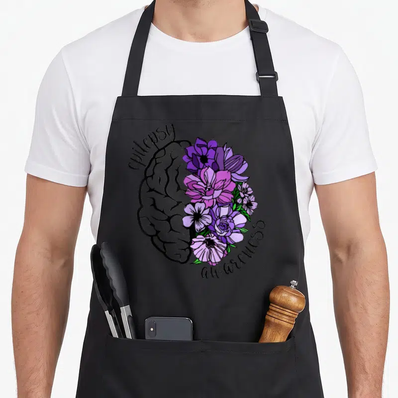 Flower epilepsy awareness apron - Split brain illustration with grey line art left side and purple pink flowers right — shown on a black cotton canvas apron against a white background