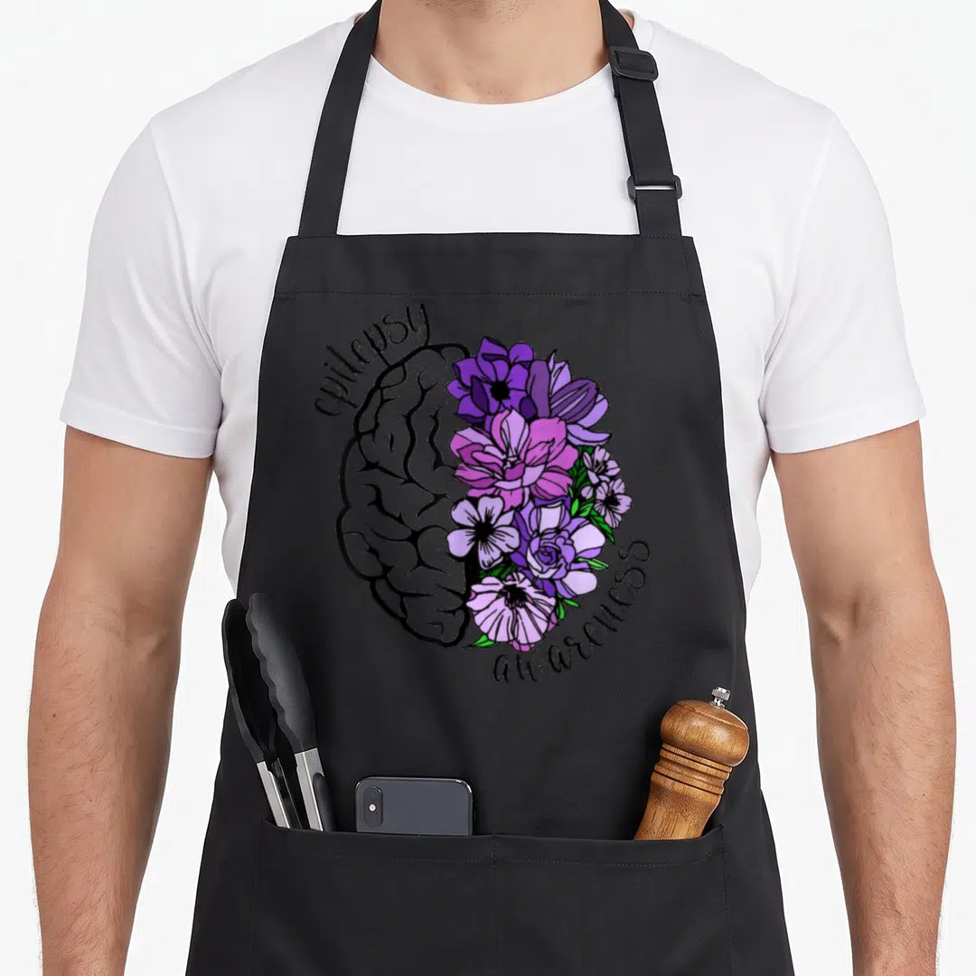 Flower epilepsy awareness apron - Split brain illustration with grey line art left side and purple pink flowers right — shown on a black cotton canvas apron against a white background