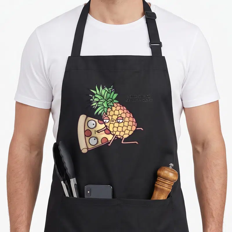 Belong pineapple apron - Cartoon pineapple hugging pizza slice with text SHHH NO ONE NEEDS TO KNOW — shown on a black cotton canvas apron against a white background