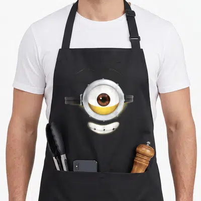 Stuart general apron - Realistic single eye with silver goggles, black eyebrows, and white smile on yellow background — shown on a black cotton canvas apron against a white background