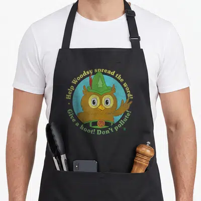 Hoot owl apron - Cartoon brown owl in green hat inside light blue circle with conservation text arching above and below — shown on a black cotton canvas apron against a white background