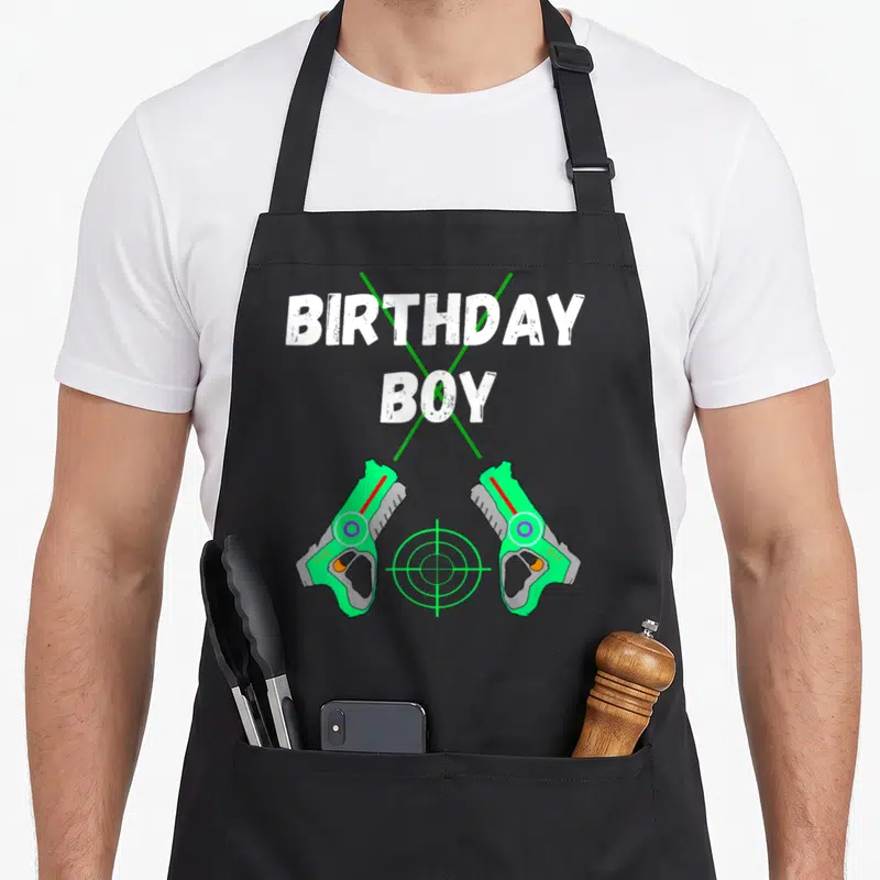 Laser tag apron - BIRTHDAY BOY text over green cross with two toy blasters and target symbol — shown on a black cotton canvas apron against a white background