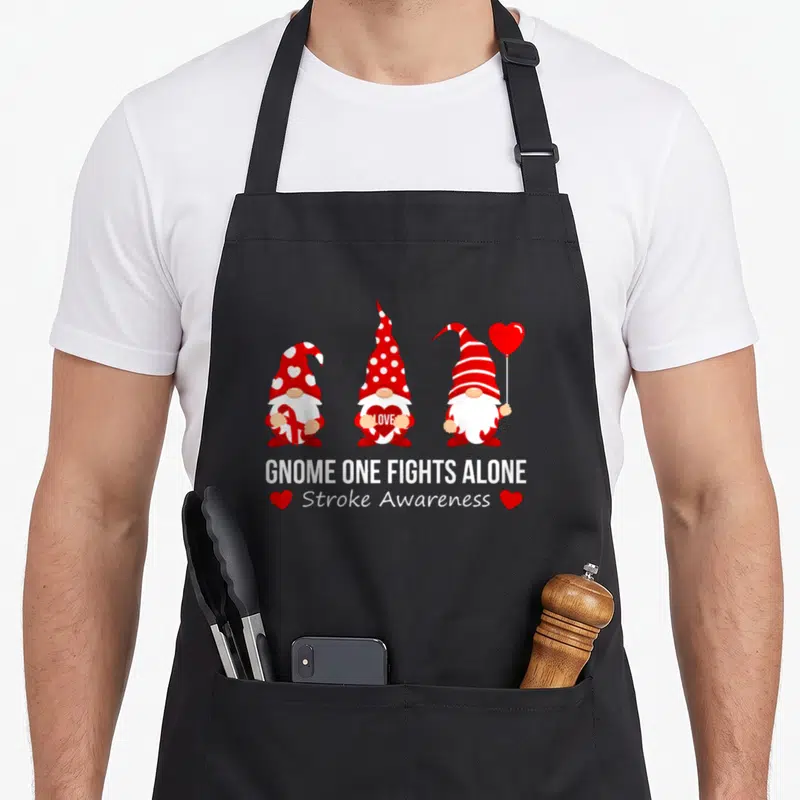Gnome fight ribbon apron - Three gnomes holding ribbon and hearts above Stroke Awareness text in red and white — shown on a black cotton canvas apron against a white background