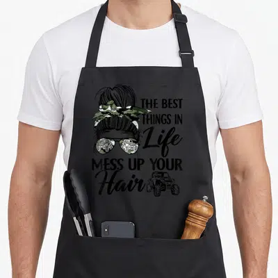 Mess utv hair apron - Woman's head with bun and camo bow, stacked text, UTV graphic, black green brown — shown on a black cotton canvas apron against a white background