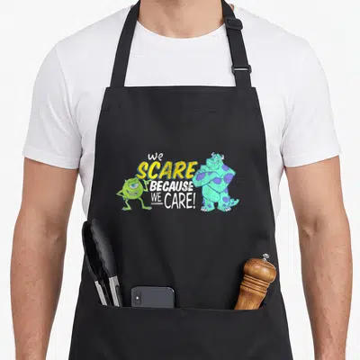 Scare panic attack apron - Sulley and Mike flanking We Scare Because We Care text in yellow and white — shown on a black cotton canvas apron against a white background