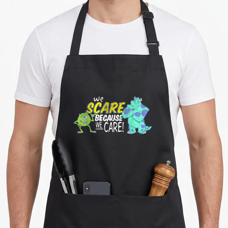 Scare panic attack apron - Sulley and Mike flanking We Scare Because We Care text in yellow and white — shown on a black cotton canvas apron against a white background