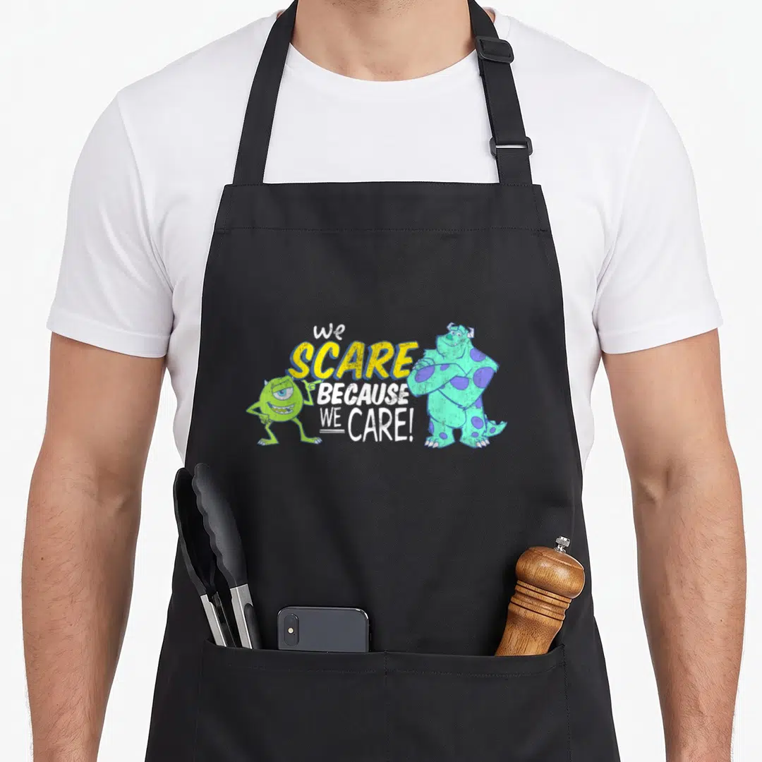 Scare panic attack apron - Sulley and Mike flanking We Scare Because We Care text in yellow and white — shown on a black cotton canvas apron against a white background