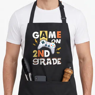 Grade game 2nd apron - GAME ON 2ND GRADE text with cartoon controller and lightning bolts — shown on a black cotton canvas apron against a white background