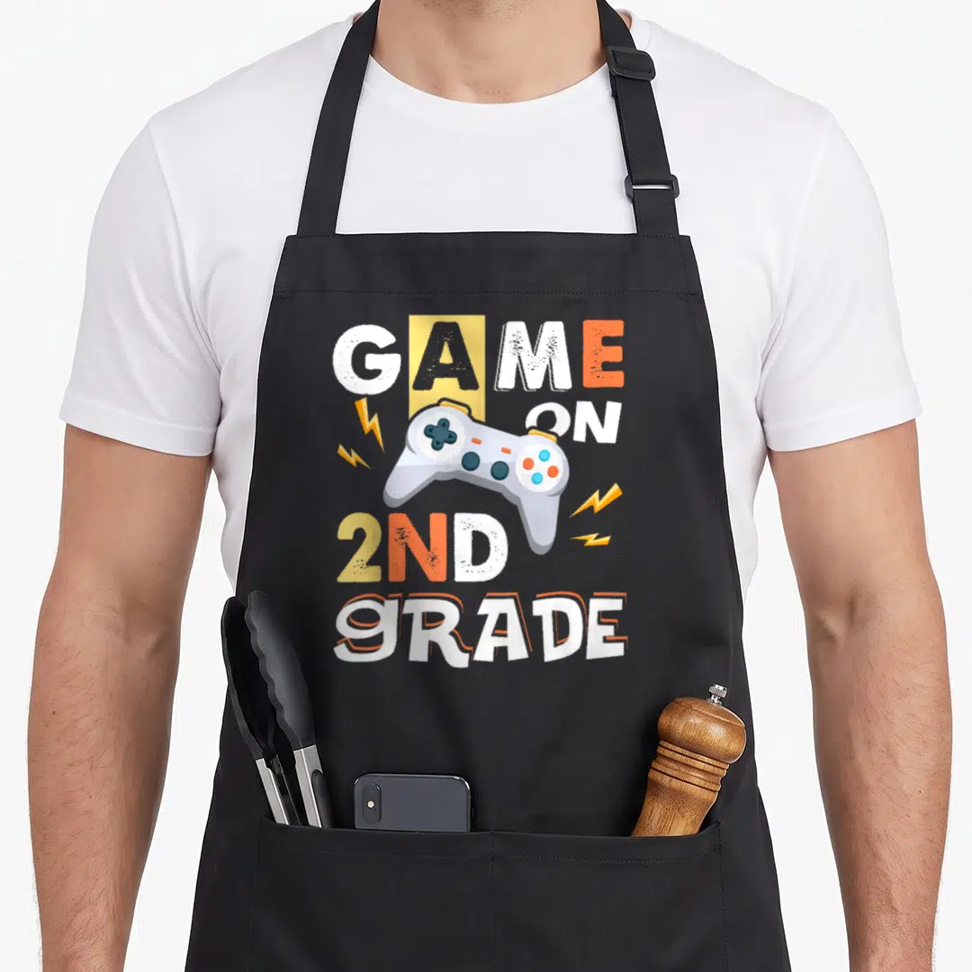 Grade game 2nd apron - GAME ON 2ND GRADE text with cartoon controller and lightning bolts — shown on a black cotton canvas apron against a white background
