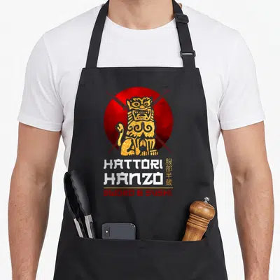 Oni fighter apron - Gold komainu on red sun with crossed swords and Hattori Hanzo Sword & Sushi text — shown on a black cotton canvas apron against a white background