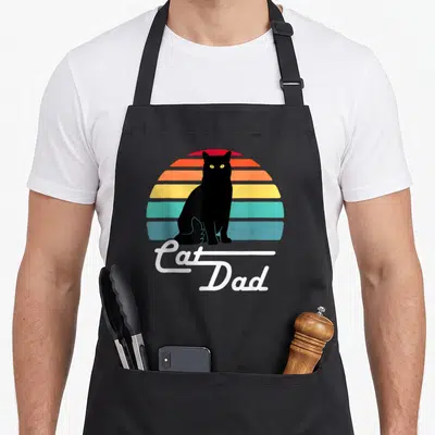 Funny cat dad apron - Black cat silhouette against retro striped sun with white Cat Dad cursive text — shown on a black cotton canvas apron against a white background