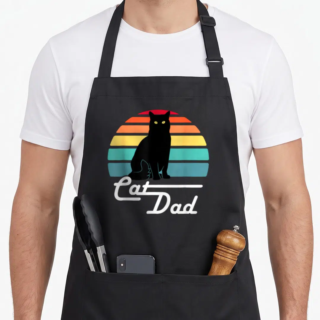 Funny cat dad apron - Black cat silhouette against retro striped sun with white Cat Dad cursive text — shown on a black cotton canvas apron against a white background