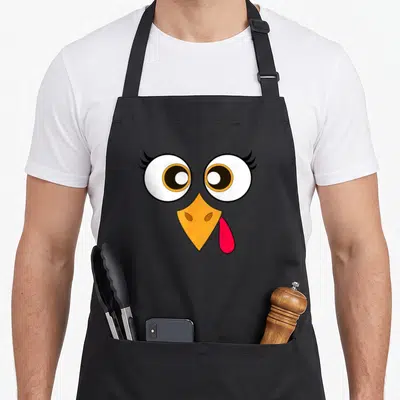 Turkey face thanksgiving day aprons - Cartoon turkey face with two large eyes, orange beak, and red wattle — shown on a black cotton canvas apron against a white background
