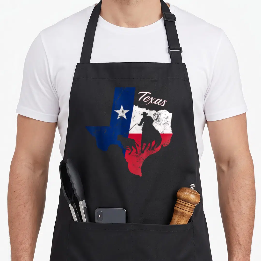 State texans apron - Distressed Texas map with cowboy silhouette and star in blue white red — shown on a black cotton canvas apron against a white background