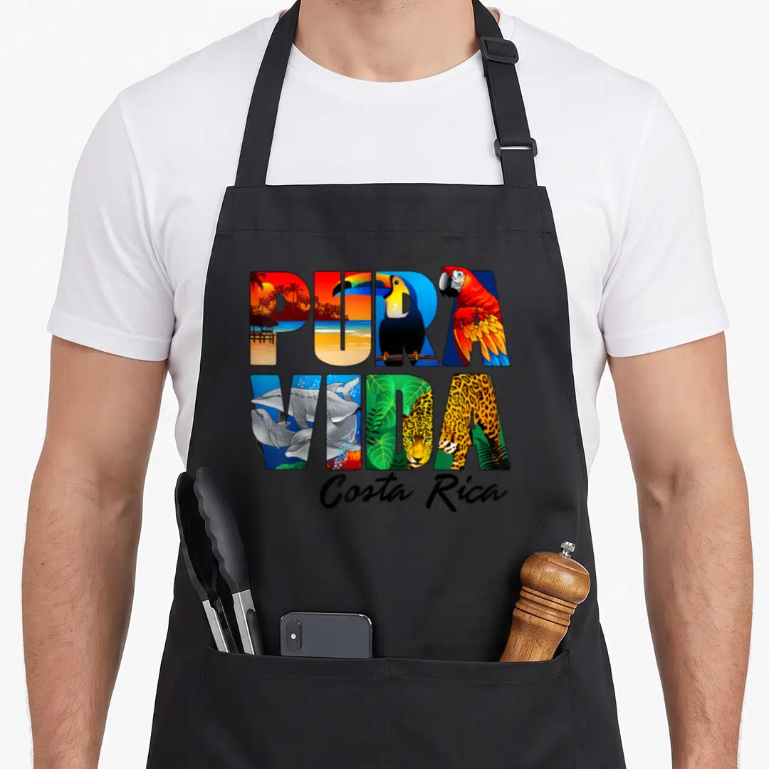 Rica costa pride apron - PURA VIDA text filled with toucan, jaguar, dolphins, and beach scenes in green blue yellow red — shown on a black cotton canvas apron against a white background