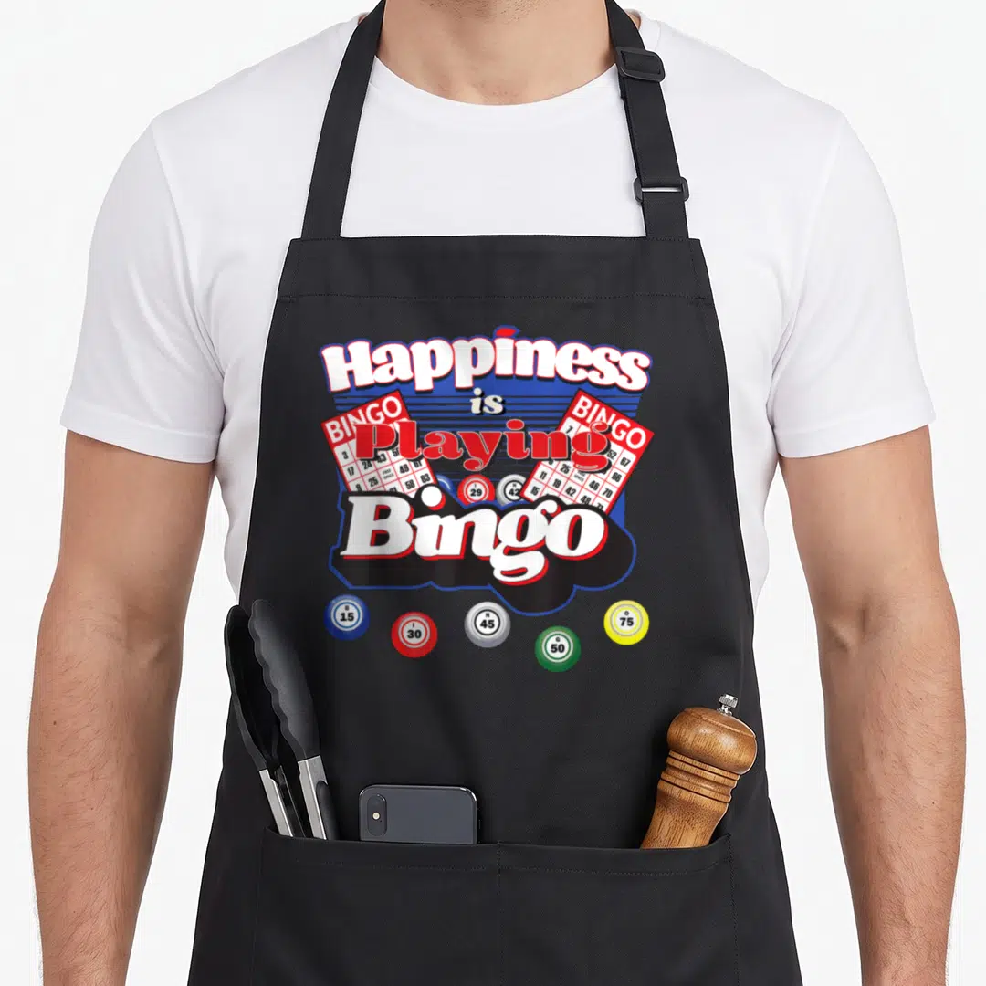 Happiness bingo apron - Happiness is Playing Bingo text with two red cards and five colored balls in red white blue — shown on a black cotton canvas apron against a white background