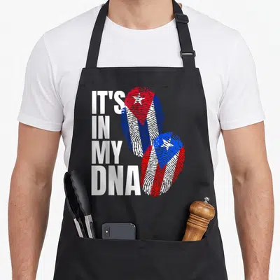 Cuban heritage aprons - IT'S IN MY DNA text next to two fingerprints filled with red white and blue flag patterns — shown on a black cotton canvas apron against a white background