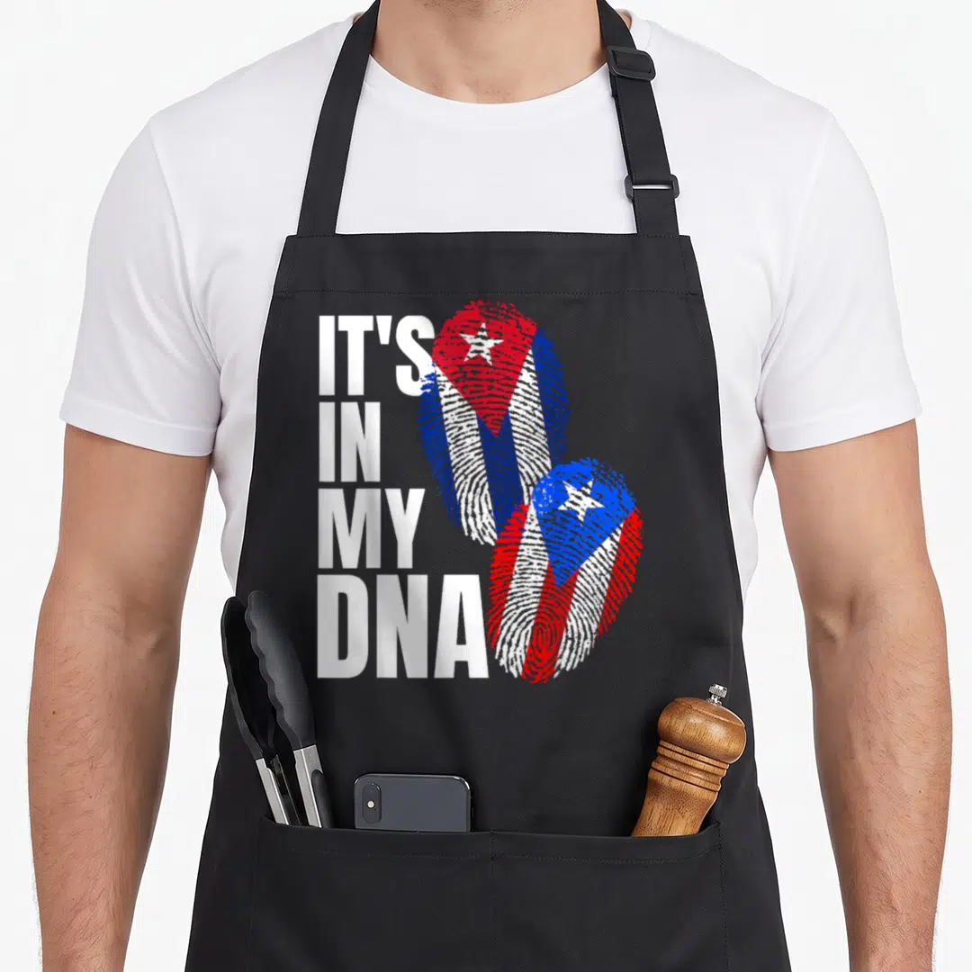 Cuban heritage aprons - IT'S IN MY DNA text next to two fingerprints filled with red white and blue flag patterns — shown on a black cotton canvas apron against a white background