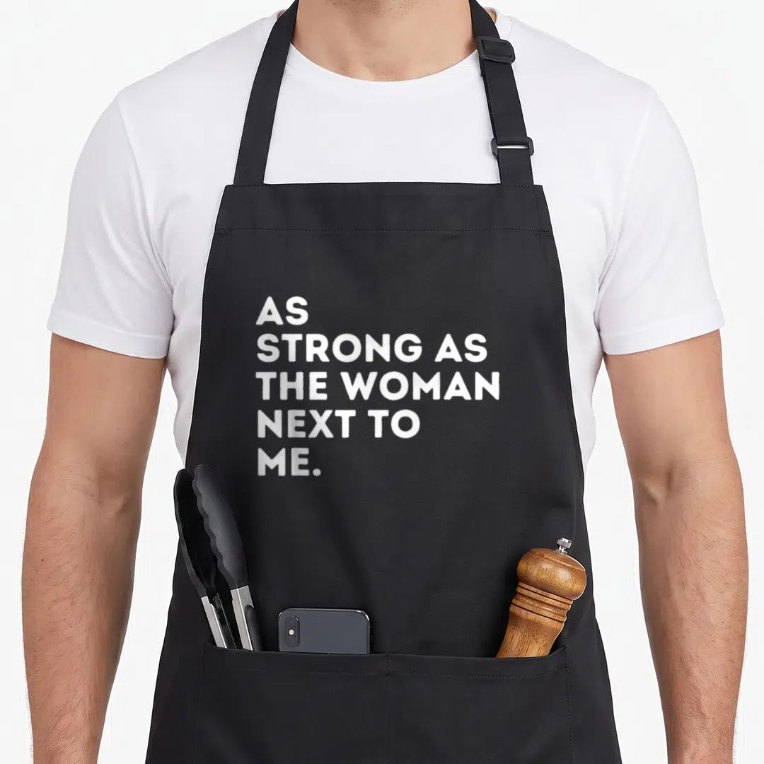 Strong a's aprons - Stacked white text AS STRONG AS THE WOMAN NEXT TO ME on black fabric — shown on a black cotton canvas apron against a white background