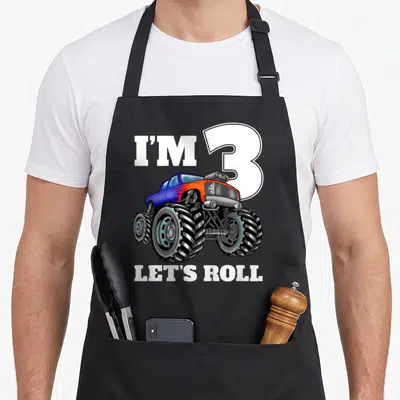Birthday three truck apron - Cartoon monster truck in blue and orange overlapping large white number 3 with text I'M 3 — shown on a black cotton canvas apron against a white background