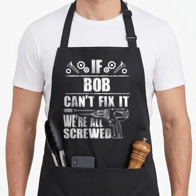 Fix birthday apron - White distressed text IF BOB CAN'T FIX IT surrounded by gears screws and a drill — shown on a black cotton canvas apron against a white background
