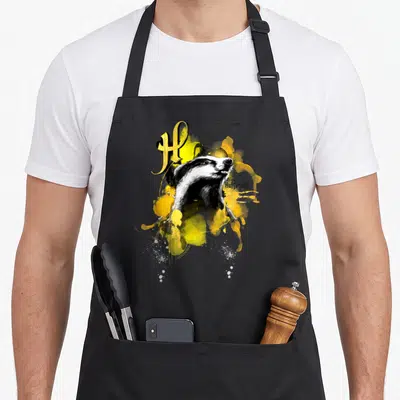 Urban badger apron - Badger illustration with Hufflepuff text and yellow paint splashes — shown on a black cotton canvas apron against a white background