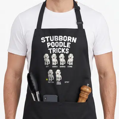 Poodle dog trainer apron - Distressed text reading STUBBORN POODLE TRICKS above two rows of white poodles and a yellow — shown on a black cotton canvas apron against a white background
