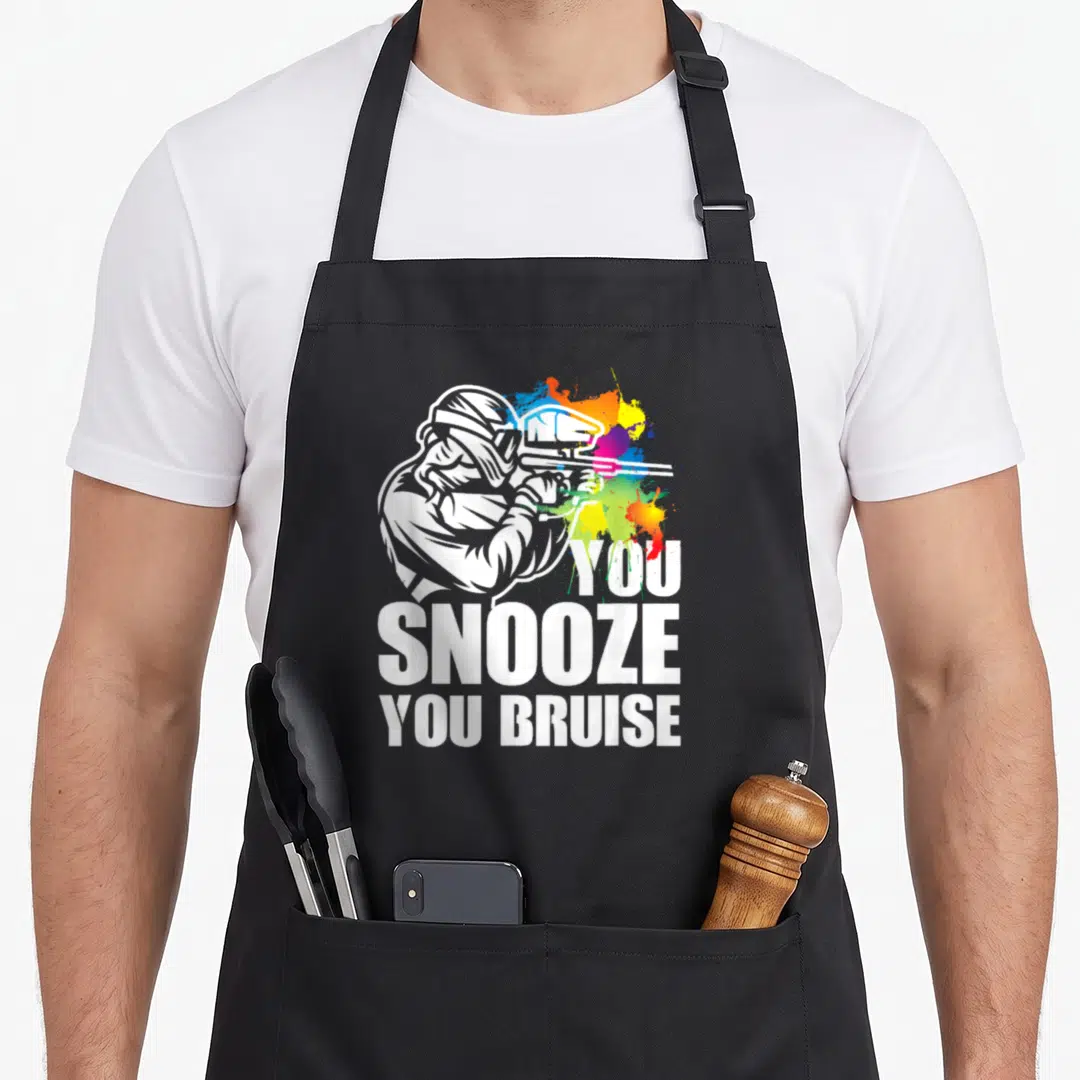 Snooze paintball apron - Cartoon paintball player aiming marker beside YOU SNOOZE YOU BRUISE text and colorful splashes — shown on a black cotton canvas apron against a white background