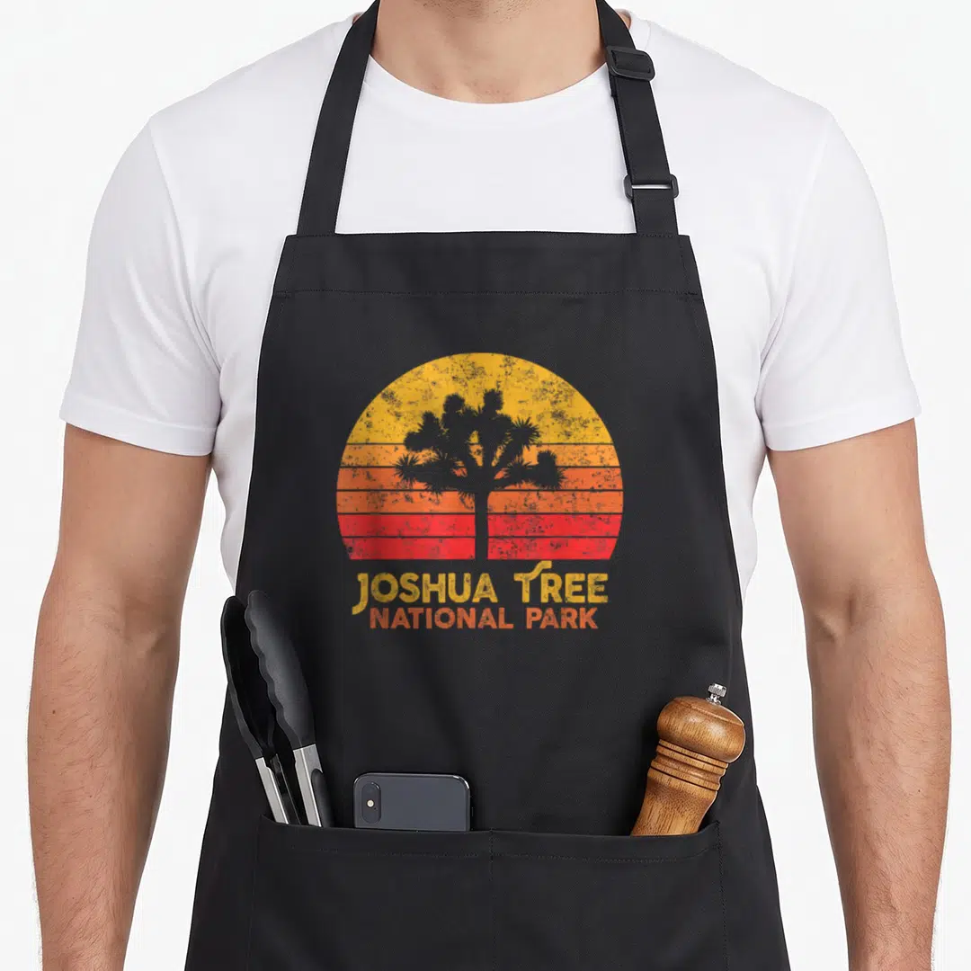 Joshua national tree apron - Grey joshua tree silhouette over yellow orange red striped sun with stacked JOSHUA TREE — shown on a black cotton canvas apron against a white background