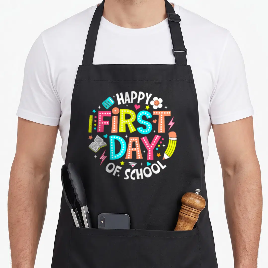 Cute student teacher apron - Circular happy first day of school text surrounded by ruler, calculator, pencil — shown on a black cotton canvas apron against a white background