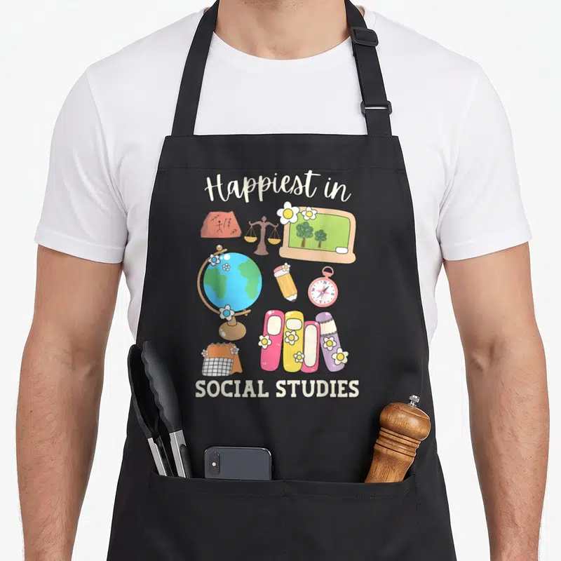 Happiest teacher apron - Cartoon social studies gear with Happiest in SOCIAL STUDIES text in beige blue pink green — shown on a black cotton canvas apron against a white background
