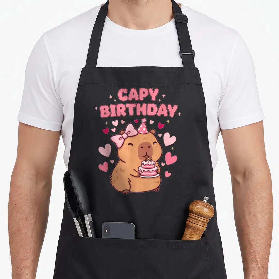 Capybara bow apron - Cartoon brown capybara holding pink cake wearing party hat and bow surrounded by hearts — shown on a black cotton canvas apron against a white background