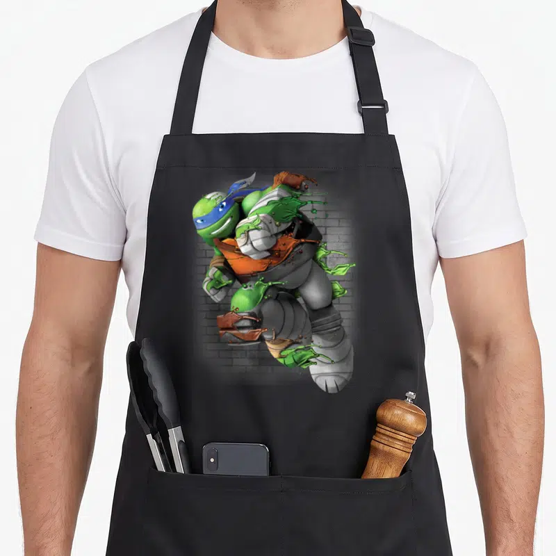 Leonardo immature apron - Realistic ninja turtle with green slime splash against grey brick wall — shown on a black cotton canvas apron against a white background