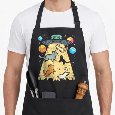 Alien space cat apron - Five cats floating in a yellow UFO tractor beam with two green aliens and planets — shown on a black cotton canvas apron against a white background