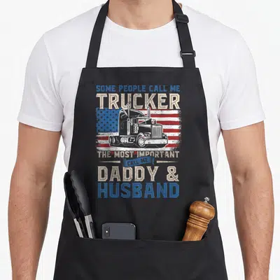 Truck american flag apron - White semi truck on distressed usa flag with daddy husband text in blue beige red white — shown on a black cotton canvas apron against a white background