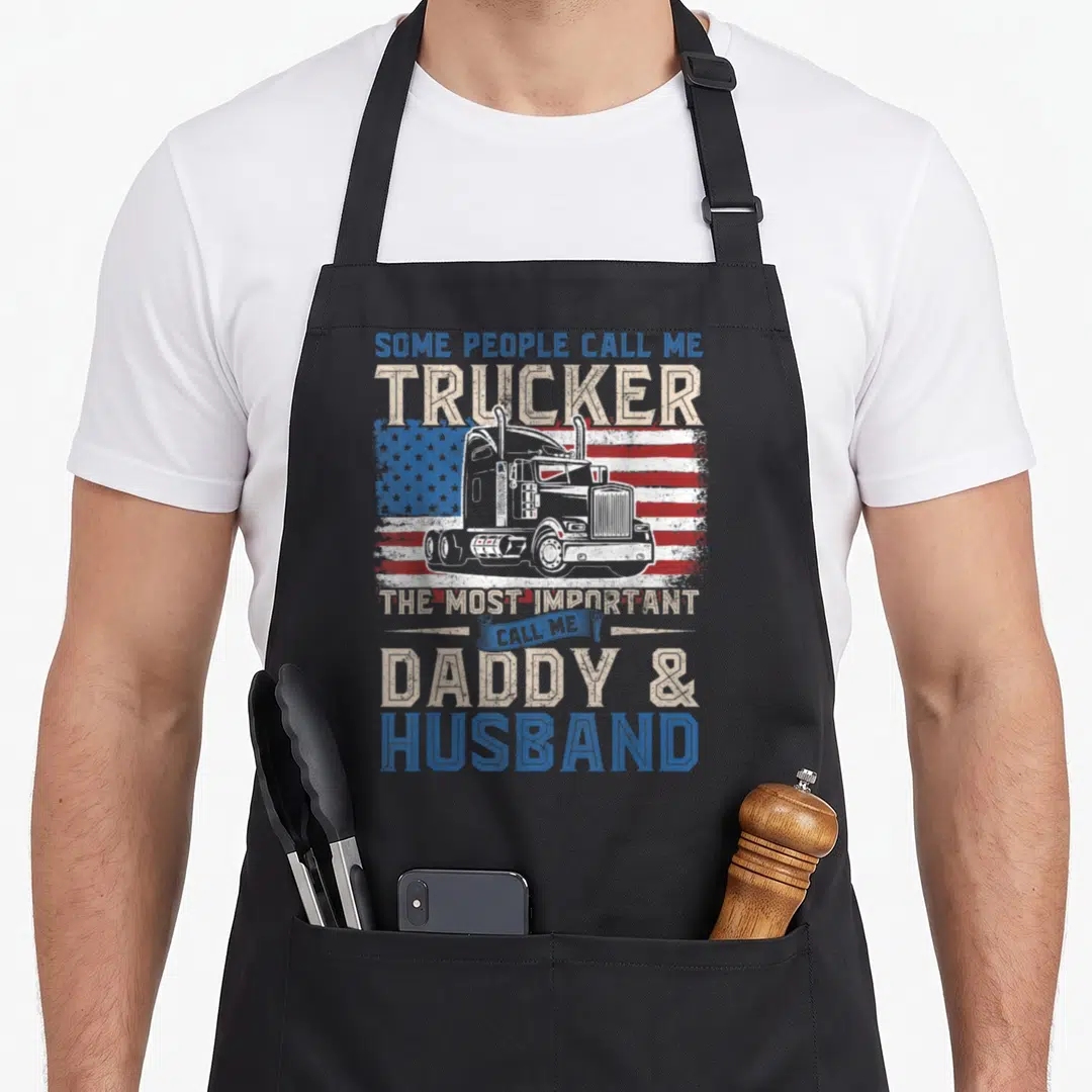Truck american flag apron - White semi truck on distressed usa flag with daddy husband text in blue beige red white — shown on a black cotton canvas apron against a white background