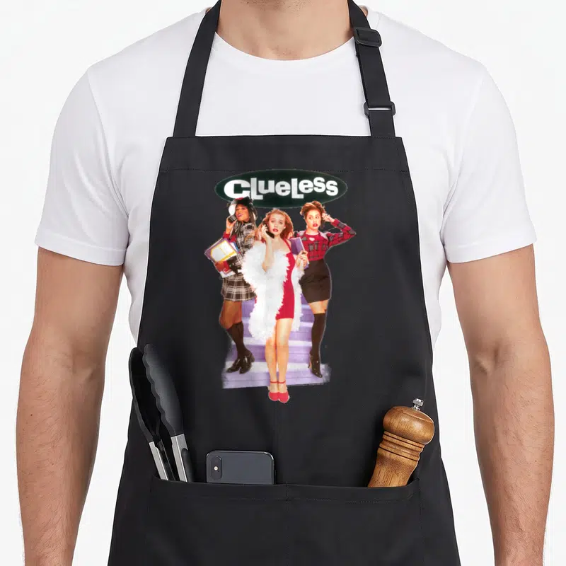 Clueless tai apron - Three women on purple stairs below white Clueless text in dark green oval — shown on a black cotton canvas apron against a white background