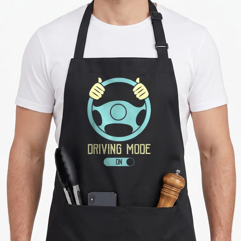 16th driving apron - Light blue steering wheel with tan hands and DRIVING MODE ON toggle switch graphic — shown on a black cotton canvas apron against a white background