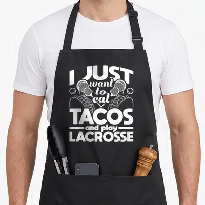 Eat taco apron - Stacked white text I JUST want to eat TACOS and play LACROSSE with sticks, balls, and tacos — shown on a black cotton canvas apron against a white background