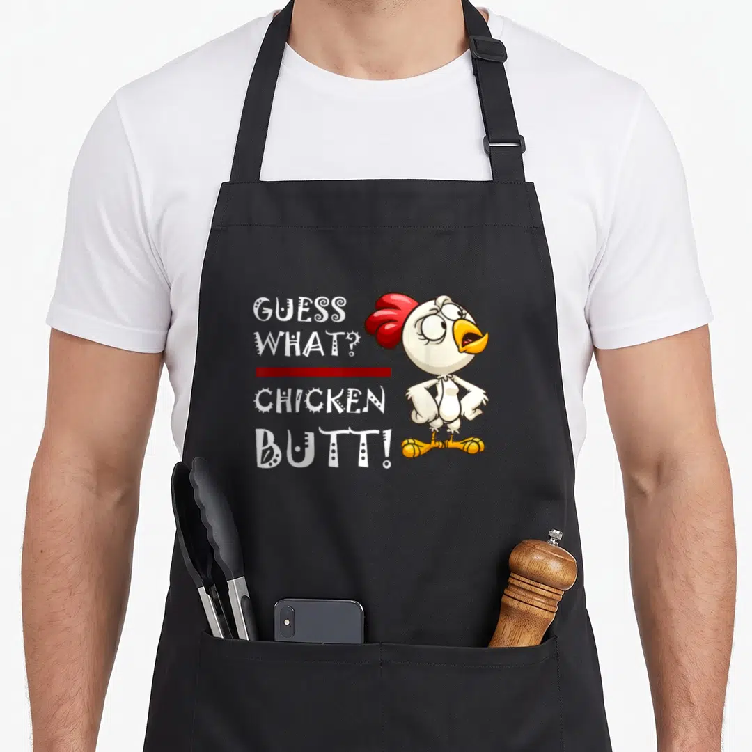 Howdy chicken coop apron - Cartoon chicken with red comb standing next to stacked white text GUESS WHAT CHICKEN BUTT — shown on a black cotton canvas apron against a white background