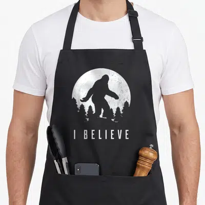 Believe sasquatch bigfoot apron - Grey Bigfoot silhouette standing before a white moon and tree line with text I — shown on a black cotton canvas apron against a white background