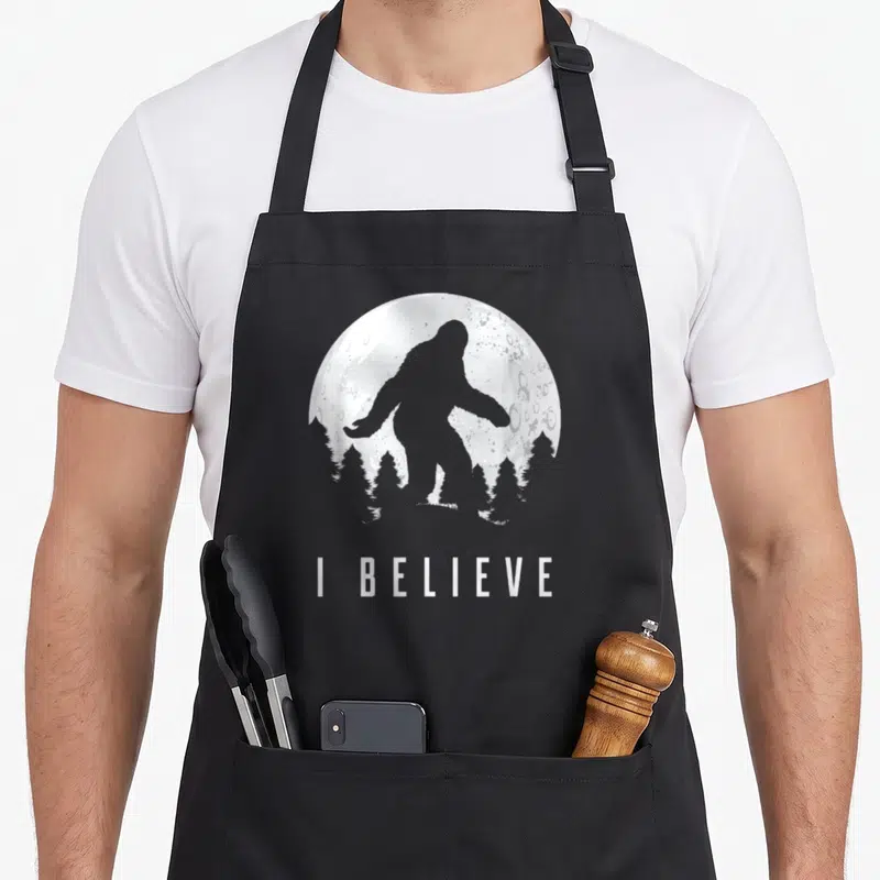 Believe sasquatch bigfoot apron - Grey Bigfoot silhouette standing before a white moon and tree line with text I — shown on a black cotton canvas apron against a white background