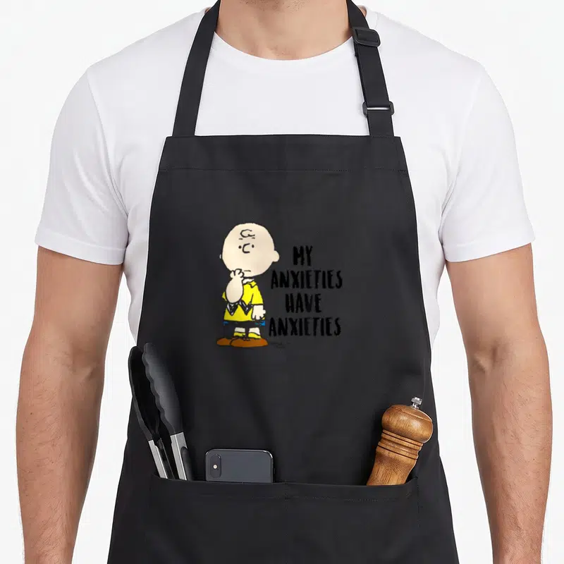 Charlie anxiety awareness apron - Cartoon boy in yellow shirt beside stacked text MY ANXIETIES HAVE ANXIETIES — shown on a black cotton canvas apron against a white background