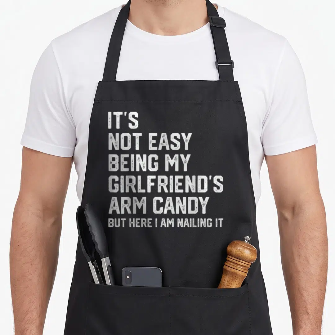 Easy father day apron - White distressed text stacked six lines reading IT'S NOT EASY BEING MY GIRLFRIEND'S ARM CANDY — shown on a black cotton canvas apron against a white background